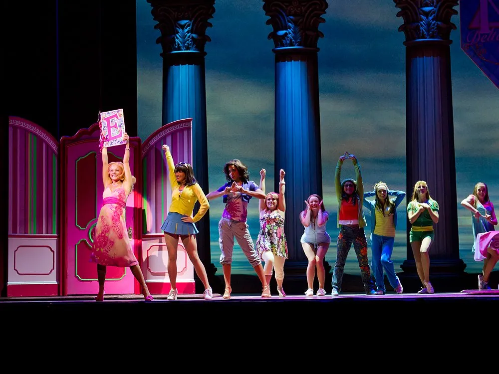 Szene: Gruppe an Darstellern in buter Bekleidung, ganz vorne Frau in rosa, hält großes "E" auf rosa Tafel