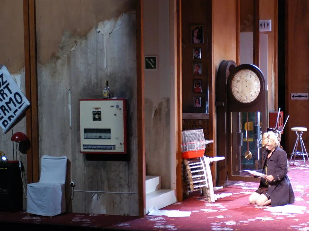 Szene: Frau sitzt am Boden , blickt auf Papier, verschiedene beschädigte Möbel im Hintergrund