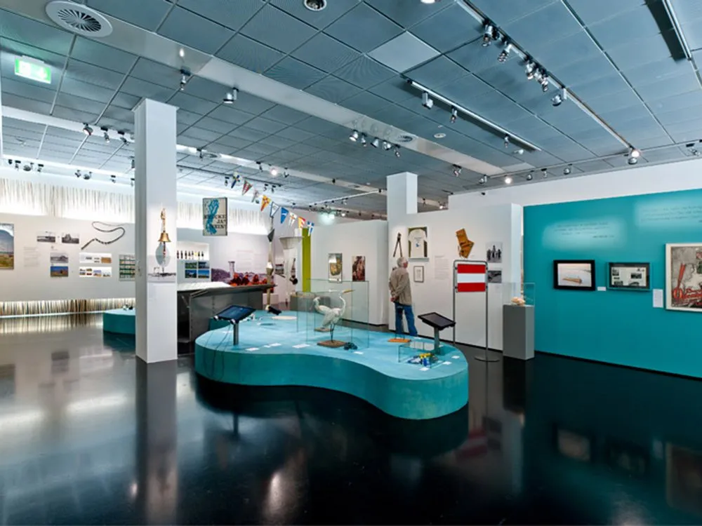 Ausstellungsraum mit einer blauen Plattform, die ein Modell eines Storches und andere Objekte zeigt, umgeben von Wänden mit Bildern, Texten und einer österreichischen Flagge
