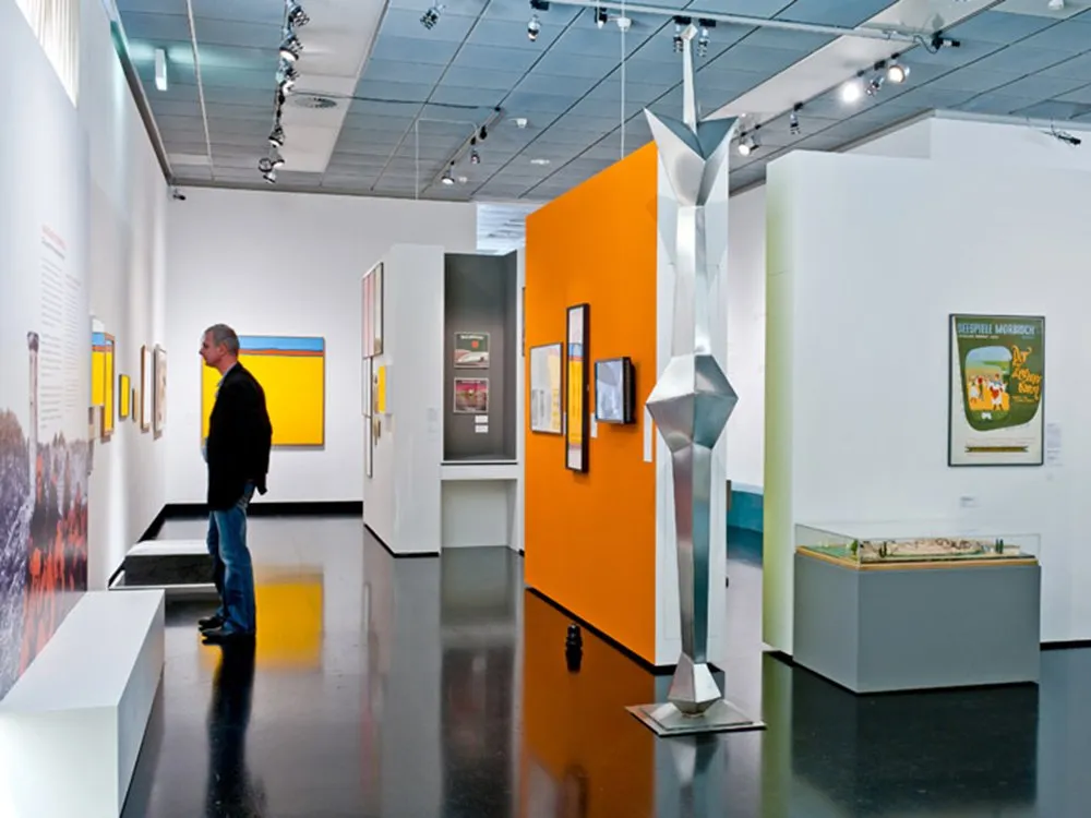 Ausstellungsraum mit einem Besucher, gelben und orangefarbenen Wänden, Kunstwerken, einer Skulptur und einem Modell in einer Vitrine