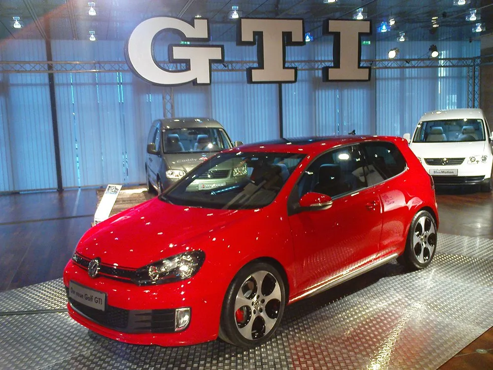 3D Schrift "GTI" hängt über rotem Auto in Ausstellungsraum 