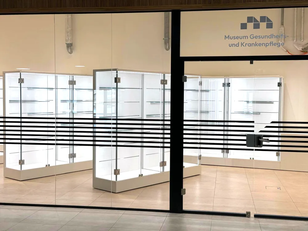 Vitrinen in einem Raum , sichtbar durch eine Glasfront mit schwarzem horizontalen Linien. Auf der Tür ein Schriftzug mit: Gesundheits- und Krankenpflege.