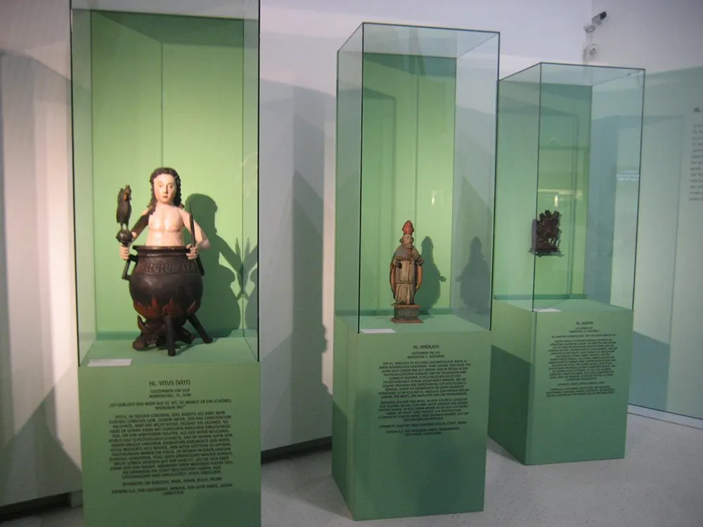 Drei Glasvitrinen mit Skulpturen, darunter Hl. Vittus in Kessel