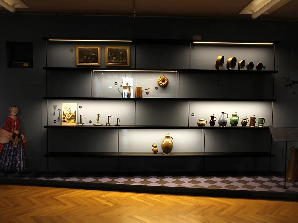 Ausstellungsraum: Schwarzes Regal mit mehreren Ebenen, gefüllt mit alten Gemälden, Kerzenständern, Krügen und einer Kuchenform. Links eine Puppe in historischer Kleidung, Boden mit Schachbrettmuster