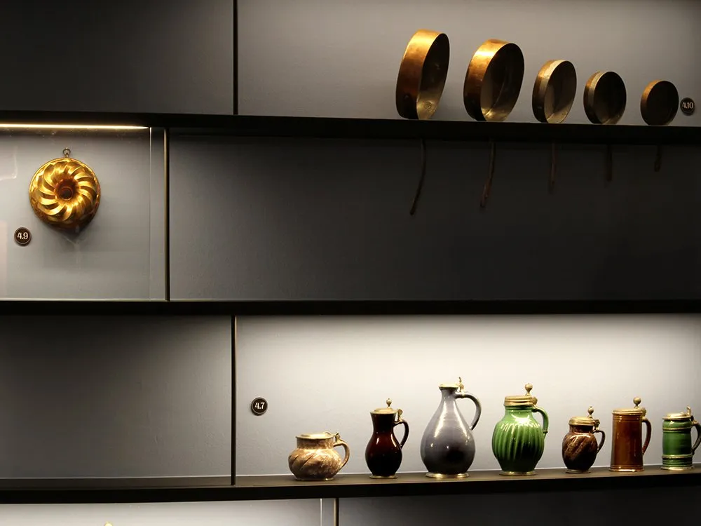 Ausstellungsraum: Schwarzes Regal mit mehreren Ebenen, gefüllt mit einer goldenen Kuchenform, Metallpfannen und verschiedenen Krügen und Bechern in unterschiedlichen Farben