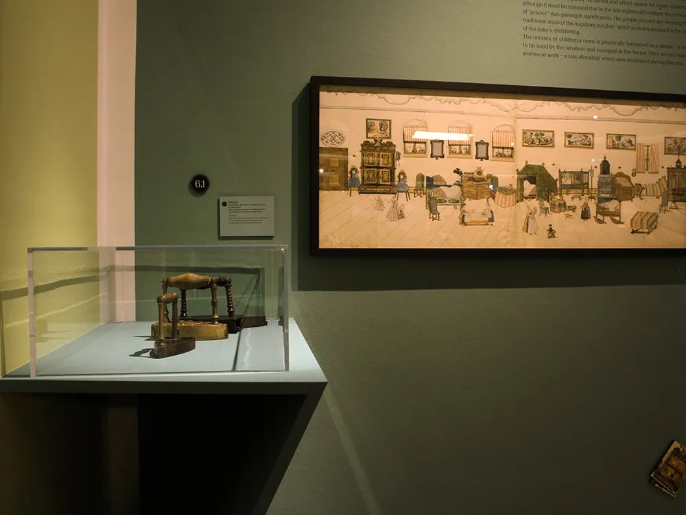 Ausstellungsraum: Vitrine mit antikem Metallgerät, daneben ein gerahmtes Bild mit Zeichnungen eines historischen Raums