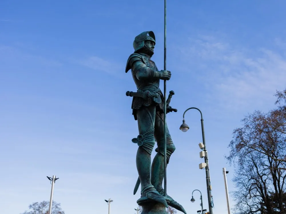 Seitenansicht der Statue, die den ganzen Körper mit Sockel zeigt. Im Hintergrund sind Straßenlaternen und Bäume zu sehen.