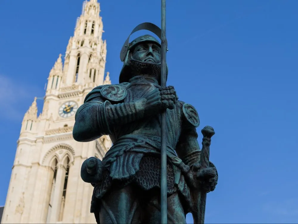 Detailansicht: Replika des Wiener Rathausmannes: Das Original ist eine 5,4 Meter hohe bronzene Ritterstatue auf der Spitze des Rathauses. Replika in Originalgröße steht vor dem Rathaus, zeigt Ritter in historischer Rüstung mit Fahne in der rechten und Schwert in der linken Hand. Im Hintergrund der Turm des Rathauses. 