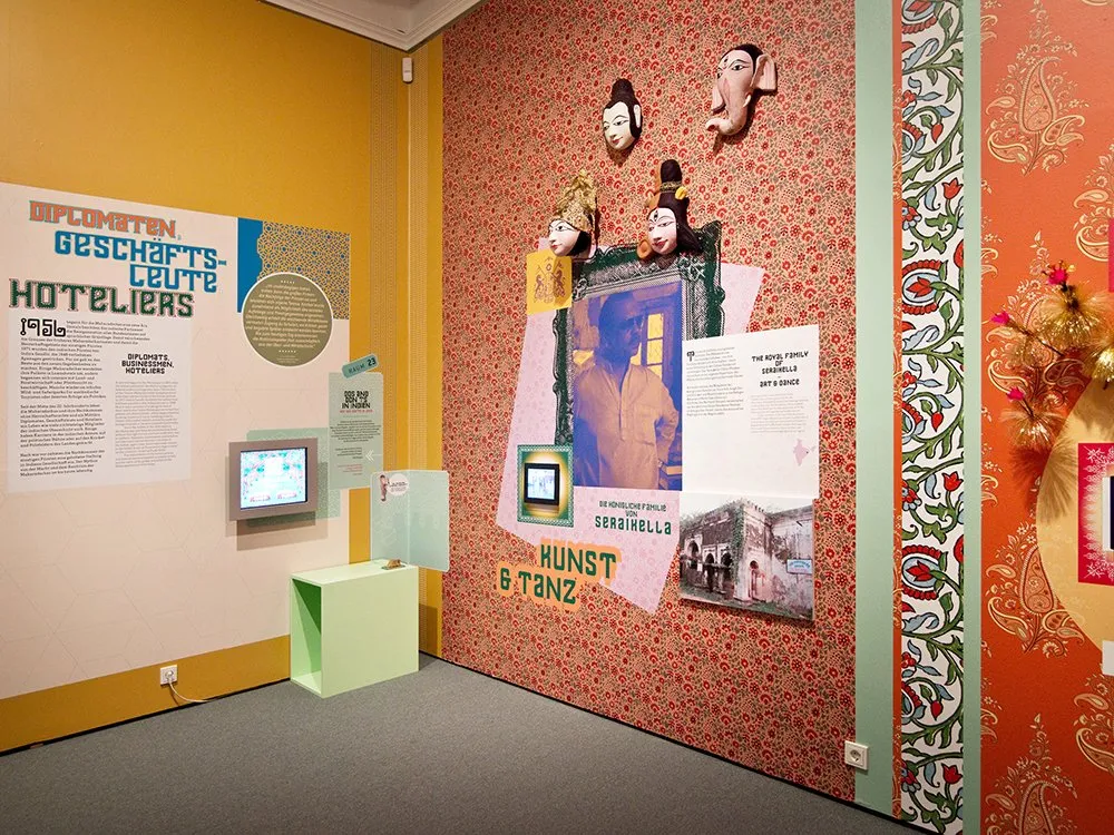 Ausstellungsraum mit orangefarbenen Wänden, verziert mit einem Paisley-Muster, und verschiedenen Ausstellungsstücken, darunter Puppenköpfe, ein Porträtfoto und informative Panels