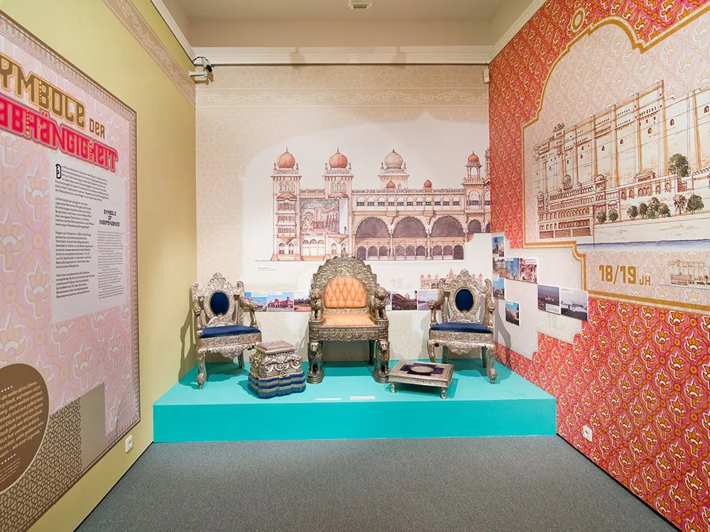 Ausstellungsraum mit einer Wandillustration eines prächtigen Palasts, drei aufwendig gestalteten Stühlen auf einer türkisfarbenen Plattform und einem Informationspanel über "Symbole der Unabhängigkeit"