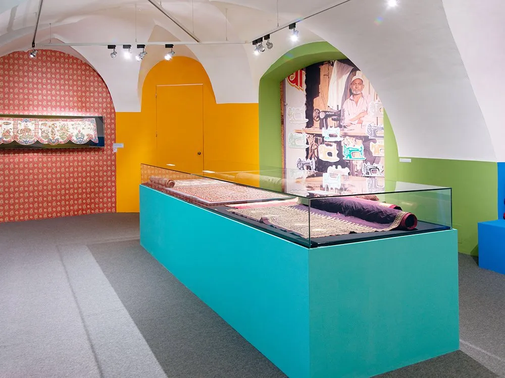 Ausstellungsraum mit gewölbter Decke, bunten Wänden in Rot, Gelb und Grün, einer Vitrine mit Teppichen und einer Wanddekoration mit einem Bild eines Mannes an einer Nähmaschine
