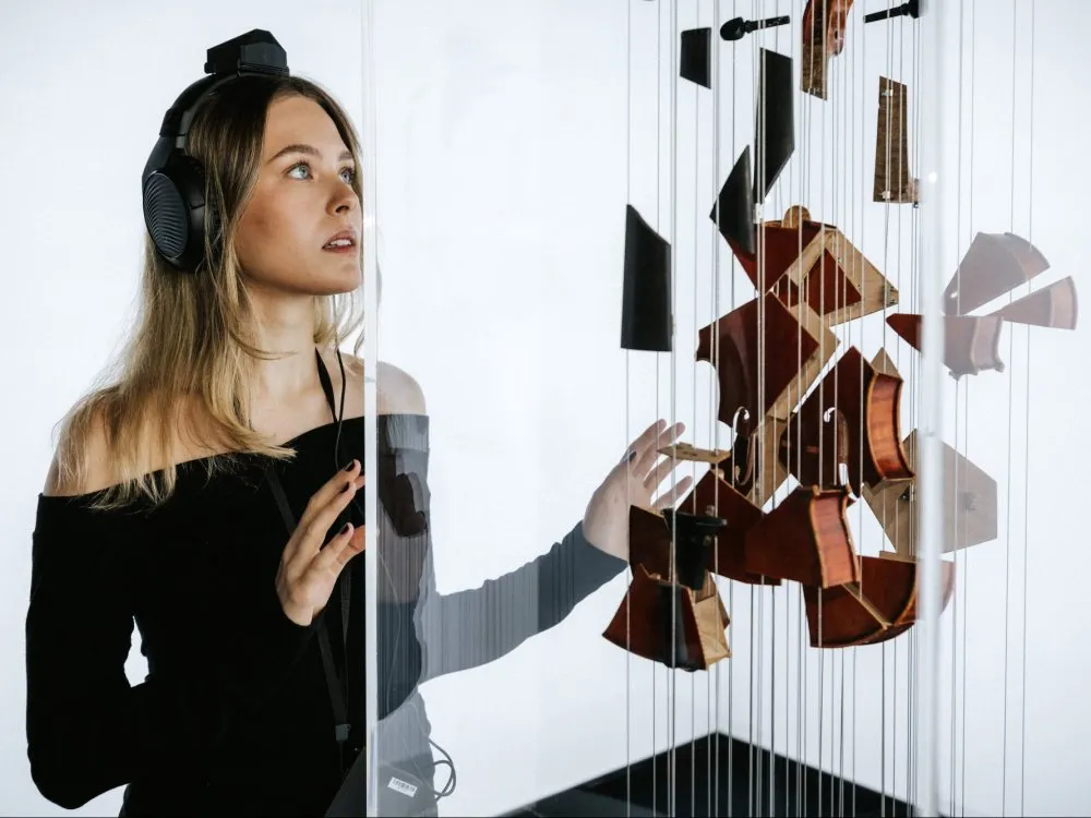 Frau mit Kopfhörern betrachtet Kunstinstallation. Vor ihr schwebt eine in Einzelteile zerschnittene Violine, deren Fragmente an dünnen Fäden hängen. Die Teile sind so ausgerichtet, dass aus einem bestimmten Blickwinkel die vollständige Form des Instruments erkennbar wird.