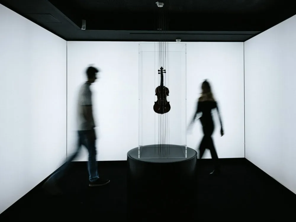 Frontalansicht der Violine-Installation aus größerer Entfernung. Zwei Personen bewegen sich um das Kunstwerk, leicht verschwommen durch Bewegungsunschärfe.