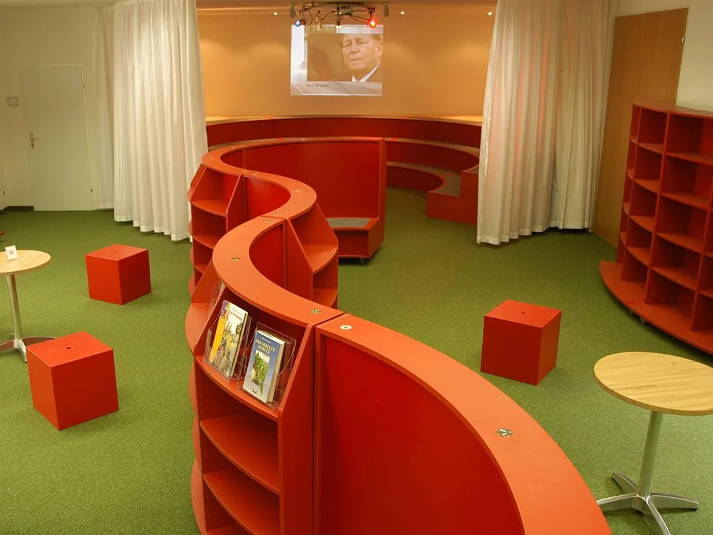 Raum mit roter, wellenförmiger Bücherregalstruktur, grünem Boden, roten Sitzwürfeln und Tischen, sowie einem projizierten Bild von Karl Ördögh an der Wand