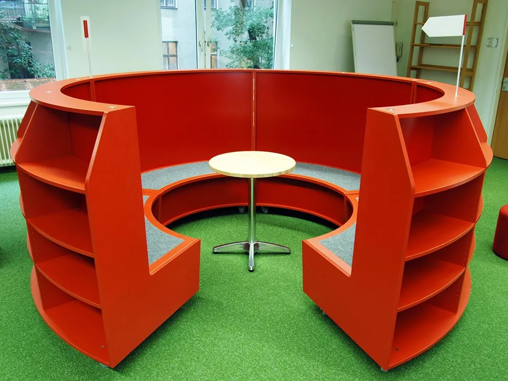 Runde rote Bücherregale mit Sitzfläche, ein runder Holztisch, roter Hocker und grüner Boden