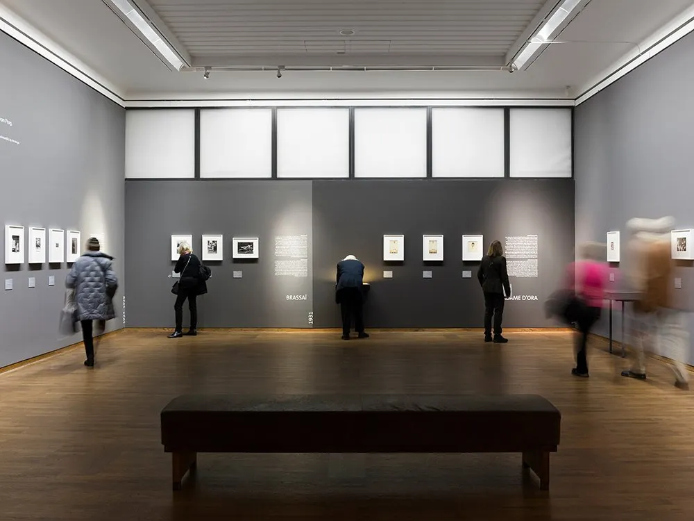 Ausstellungsraum: einige kleine Bilder an grauen Wänden, einige Besucher im Raum