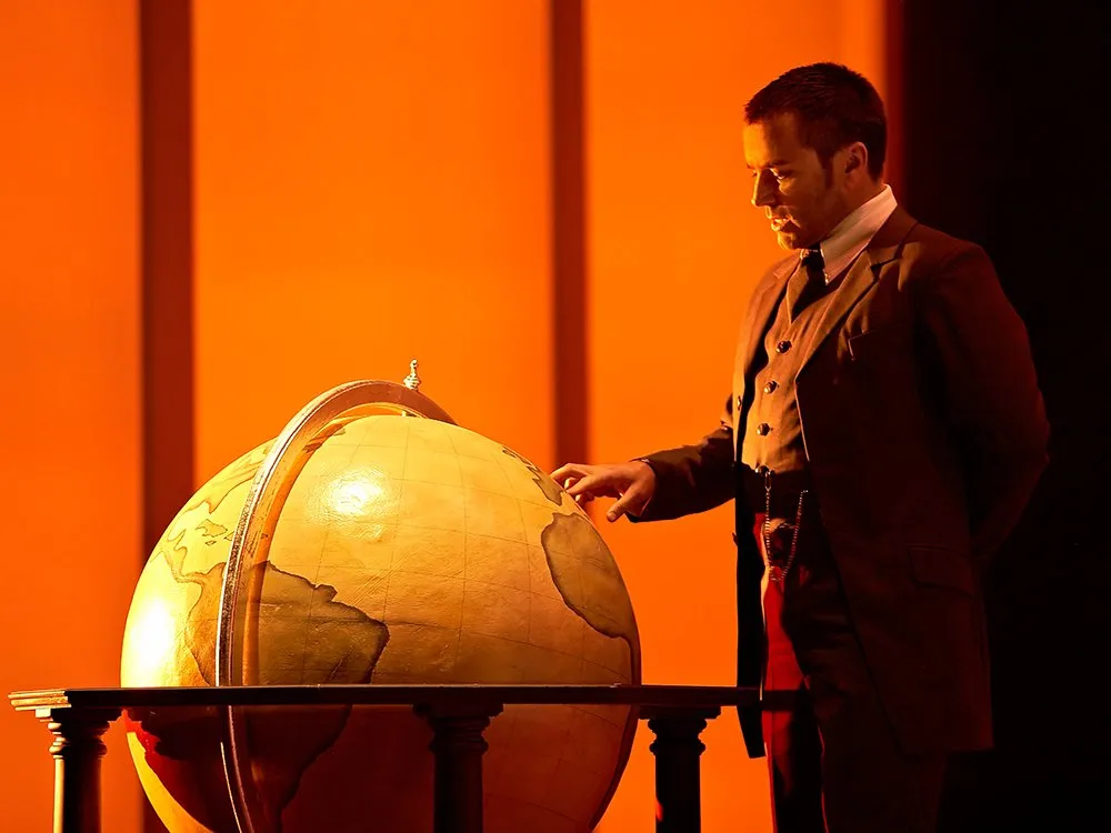 Szene: Mann in historischem Anzug steht neben einer großen, beleuchteten Globus und zeigt darauf, vor orangem Hintergrund