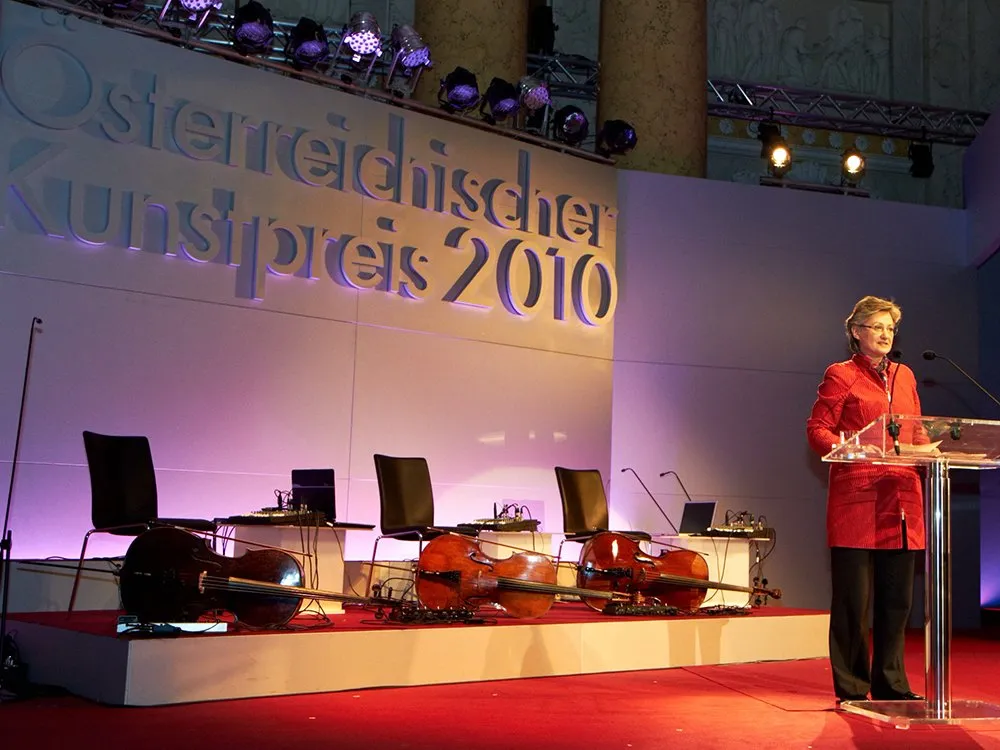3D Schrift "Österreichischer Kunstpreis 2010" an Bühnenrückwand, Frau an Pult, Streichinstrumente neben Sessel am Boden