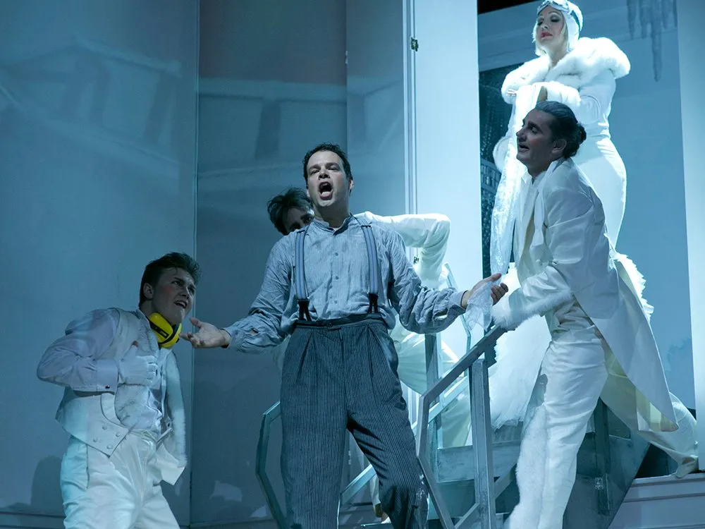 Szene: Mann in grauer Hose und Hosenträgern steht auf Treppe und singt, umringt von Darstellern in weißen Kostümen