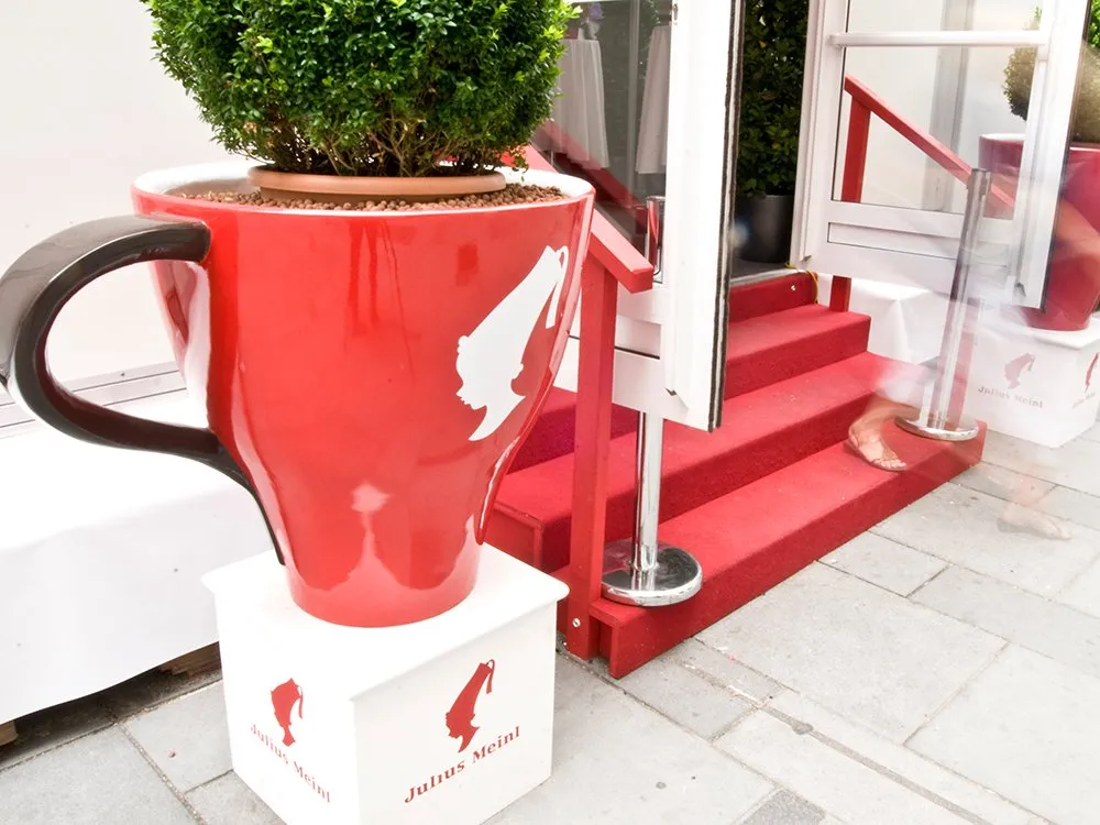 Überdimensionale rote Kaffeetasse mit Julius Meindl Logo, als Blumentopf mit Pflanze