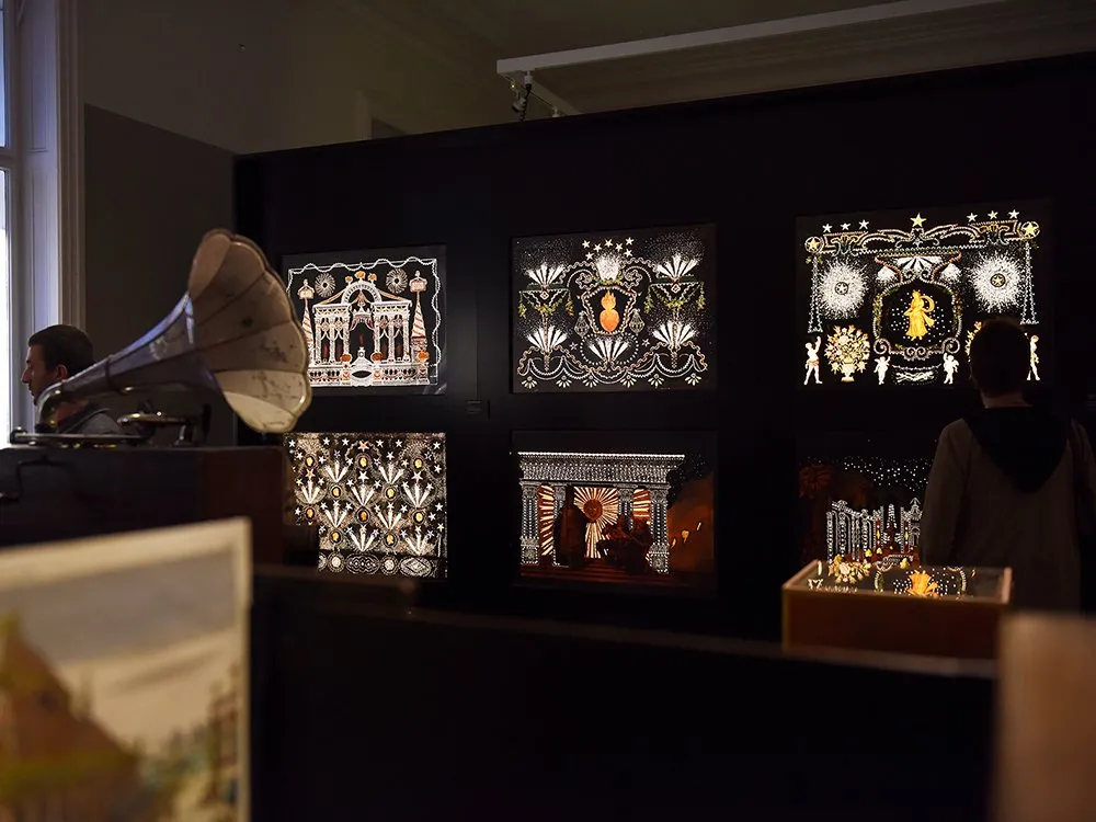 Ausstellung mit mehreren beleuchteten Kunstwerken an einer schwarzen Wand, darunter ein Grammophon und detaillierte, ornamentale Designs