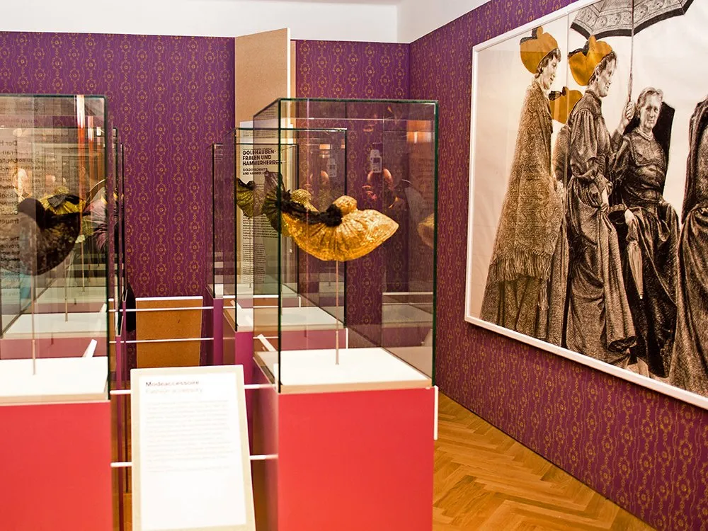 Ausstellungsraum: Vitrinen mit goldenen Modeaccessoires, darunter ein auffälliger Hut, stehen vor purpurfarbenen Wänden mit Ornamenten