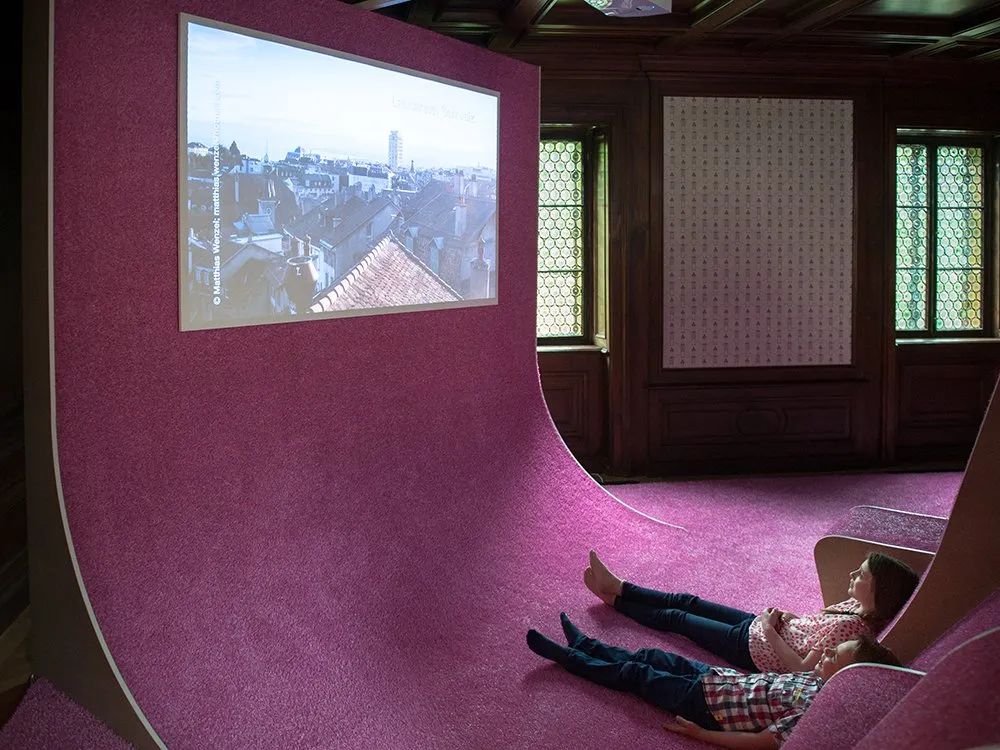 Ausstellungsraum: Zwei Kinder liegen auf einem pinken Teppich und schauen zu einer Projektion einer Stadtlandschaft
