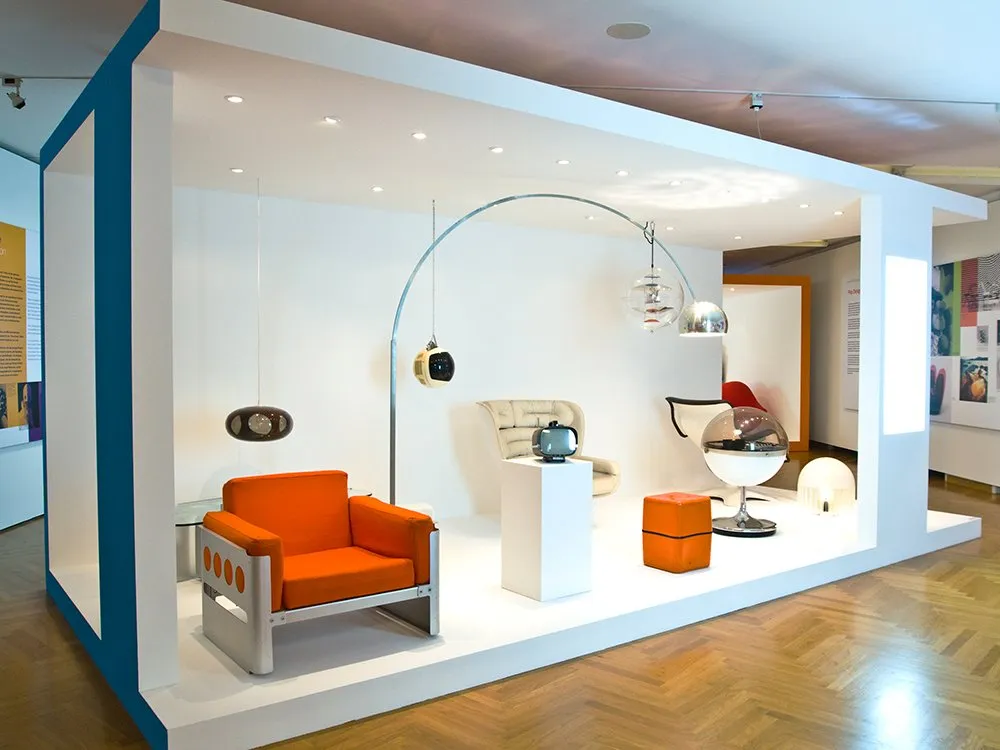 Ausstellung mit einem orangefarbenen Sessel, Retro-Möbeln, Lampen und einem Fernseher auf einem Podest