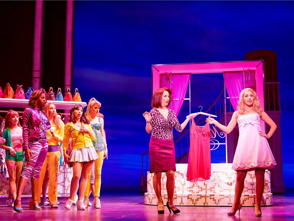 Szene: Frau in pinkem Kleid auf der Bühne, zweite in leopardengefleckter Bluse mit rosa Kleid, dahinter Frauen in bunten Outfits