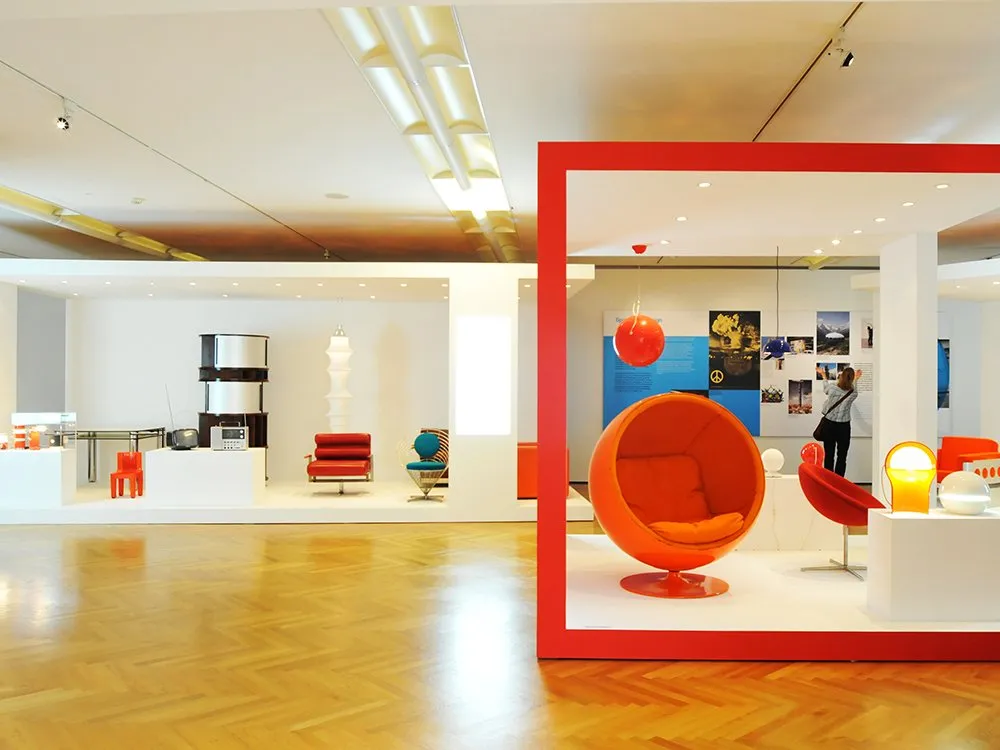 Ausstellungsraum: Moderne Ausstellung mit farbenfrohen Stühlen, darunter ein auffälliger orangefarbener Kugelsessel, sowie Retro-Möbel und historische Bilder