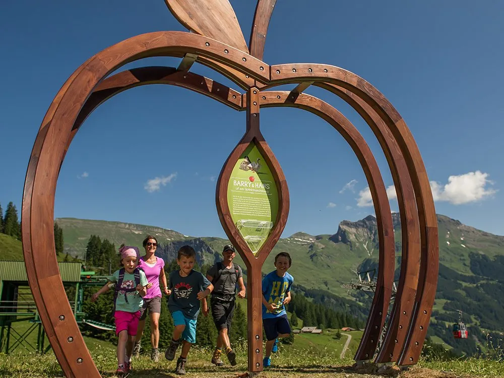 Kinder und Erwachsene laufen unter einem hölzernen Apfelbogen mit Bergen im Hintergrund