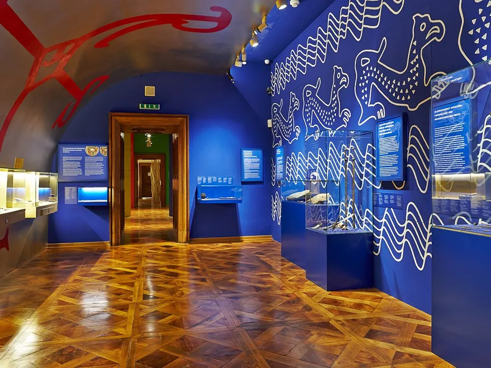 Ausstellungsraum mit blauen Wänden mit weißen Ornamenten und Vitrinen mit Artefakte