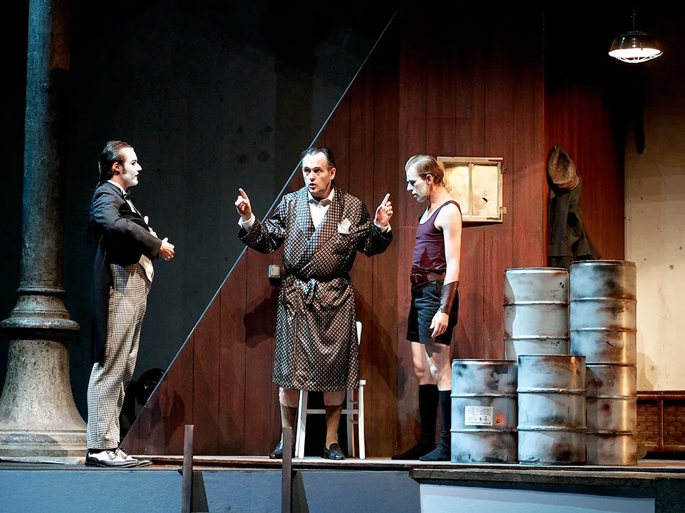 Szene: Zwei Männer mit weißer Gesichtsbemalung, einer in Anzug und der andere in Kimono, stehen neben einem dritten Mann in ungewöhnlicher Kleidung