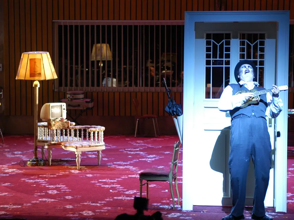 Szene: Mann steht mit Ukulele vor freistehnder Tür, roter Teppich mit alten Sesseln und Lampe im Hintergrund