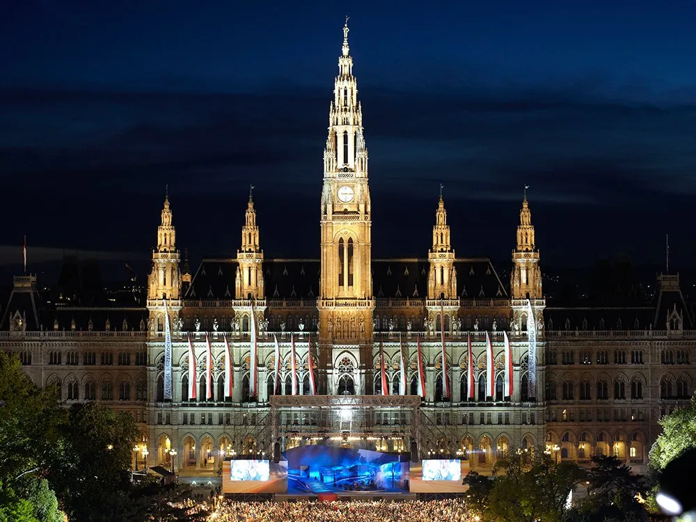 Wiener Rathaus bei Nacht, beleuchtet mit einer großen Menschenmenge und einer Bühne davor, umgeben von Flaggen und Bäumen