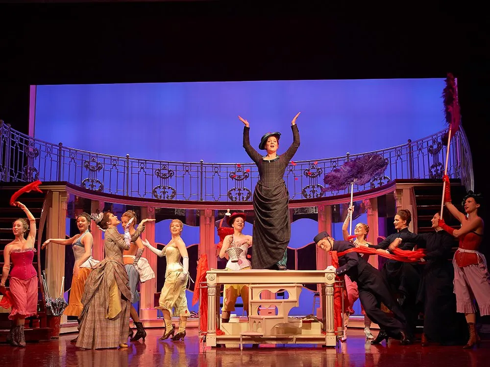 Szene: Frau steht auf einem Tisch mit erhobenen Armen, umgeben von tanzenden Darstellern  