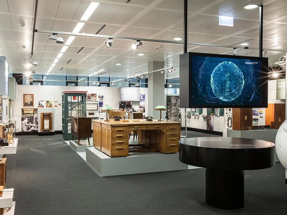 Ausstellungsraum mit historischem Büroequipment, darunter ein hölzerner Schreibtisch, alte Telefone und Radios, sowie einem modernen Bildschirm mit einem digitalen Globus im Hintergrund