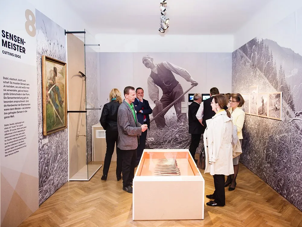 Ausstellungsraum: Personen betrachten Gemälde, ein Foto eines Mannes mit Sichel und eine Vitrine mit Werkzeugen, umgeben von bergiger Wanddekoration