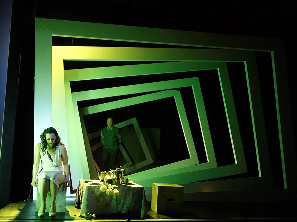 Szene: Frau in Weiß an einem Tisch mit Blumen und Getränken, einem Mann im Hintergrund, umgeben von grünen, verzerrten Rahmen