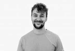 Halbporträt in schwarz-weiß: Mann mit lockigen Haaren und Bart, trägt helles Oberteil