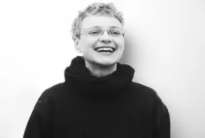 Halbporträt in schwarz-weiß: Frau mit kurzen, lockigen Haaren und Brille, trägt dunkles Oberteil