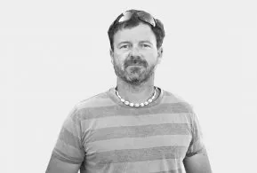 Halbporträt in schwarz-weiß: Mann mit kurzen Haaren und Bart, trägt gestreiftes Oberteil mit Halskette