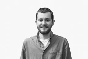 Halbporträt in schwarz-weiß: lächelnder Mann mit kurzem Bart und kurzen Haaren, trägt Hemd über T-Shirt 