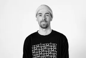 Halbporträt in schwarz-weiß: Mann mit Mütze und Bart, trägt bedrucktes dunkles Oberteil