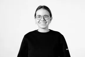 Halbporträt in schwarz-weiß: lächelnde Frau mit Brille und zusammengebundenen Haaren, trägt dunkles Oberteil