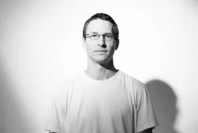 Halbporträt in schwarz-weiß: Mann mit kurzen Haaren und Brille, trägt helles T-Shirt