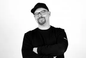 Halbporträt in schwarz-weiß: Mann mit Brille und Kappe, trägt dunkles Oberteil und hat Arme verschränkt