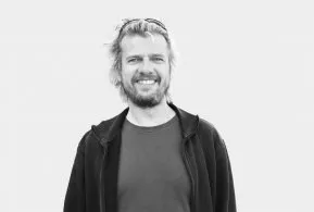 Halbporträt in schwarz-weiß: Mann mit langen Haaren und Bart, trägt dunkle Jacke über dunklem Oberteil