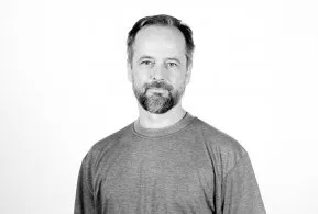 Halbporträt in schwarz-weiß: Mann mit kurzen Haaren und Bart, trägt helles Oberteil