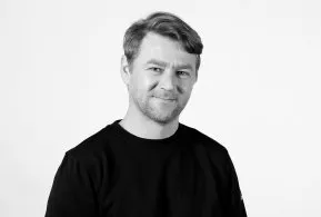 Halbporträt in schwarz-weiß: Mann mit kurzen Haaren und Bart, trägt dunkles Oberteil